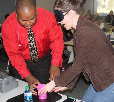 Walter Blackmon showing a woman wearing a blindfold how someone who is blind pours liquid into a glass without overflowing it.